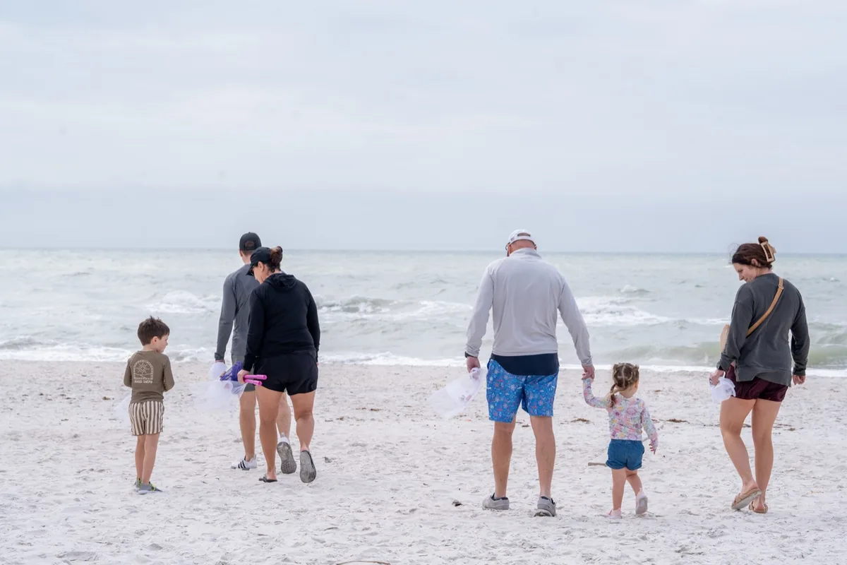 A family walks a wide white-sand beach carrying shell bags on a cloudy day